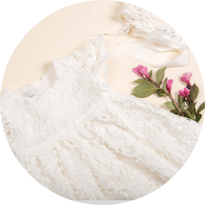 A delicate white lace baby dress is pictured alongside matching lace accessories. Adjacent to the dress are sprigs of pink flowers with green leaves, all arranged on a soft cream-colored background.