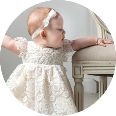 A baby in a white lace dress and headband stands while holding onto a chair.