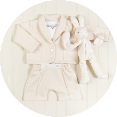 A cream-colored baby outfit with a jacket and pants is laid out on a light wood surface. A matching plush bunny toy with long ears is placed beside the outfit. The set has a soft and cozy appearance.