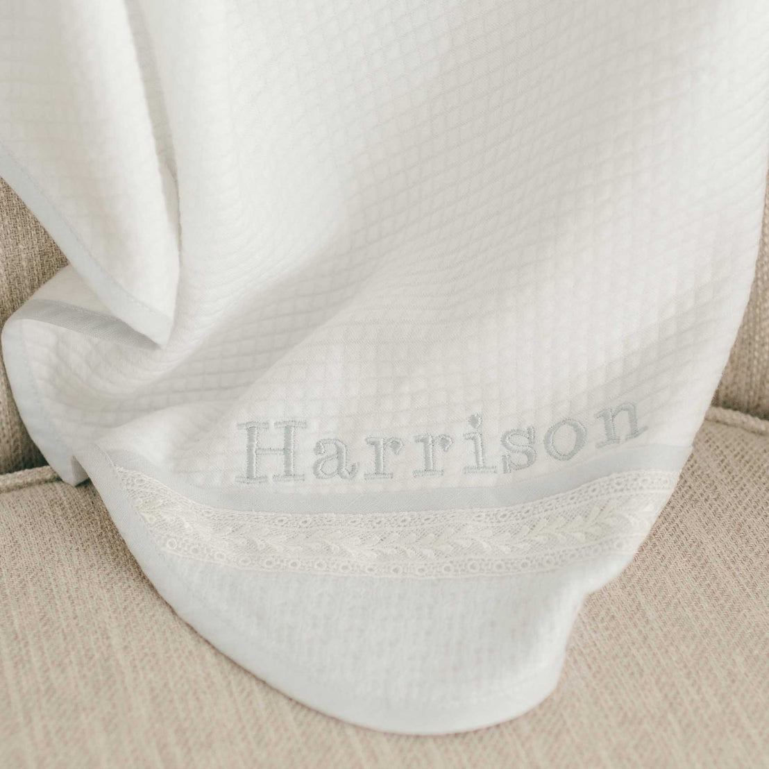 Close-up of a baby's hand gently placed on a white quilted christening blanket, personalized with "Harrison" embroidery and lace trim.