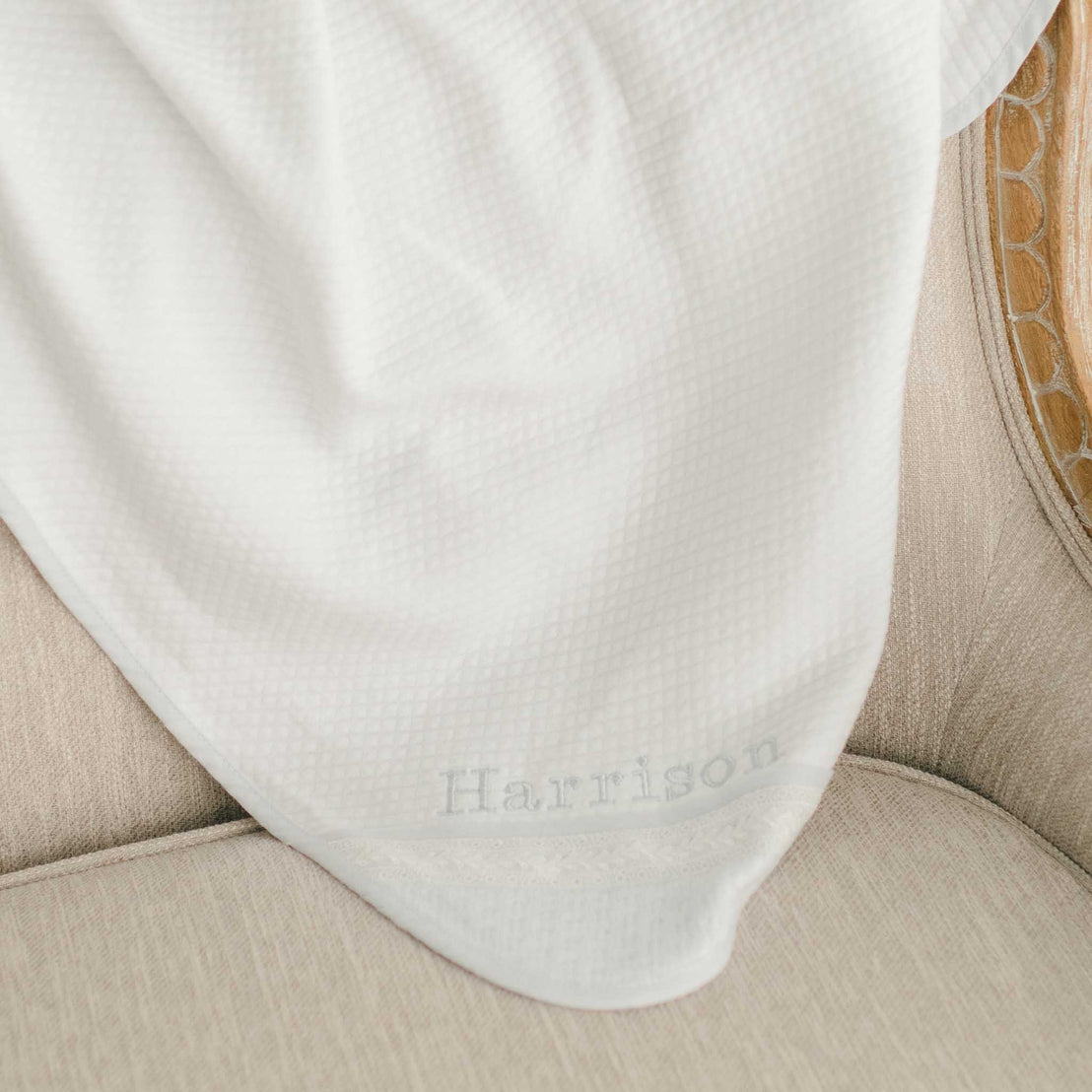 Flat lay photo the Harrison Personalized Blanket made with a plush quilted white cotton and trimmed with light blue linen. The accent corner features ivory Venice lace. On the corner of the blanket is the name "Harrison" embroidered in light blue thread