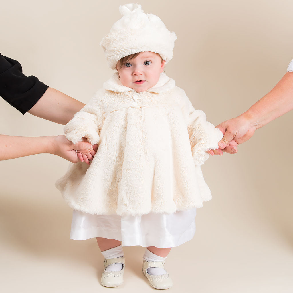 A baby is dressed in the luxurious Glad Dreams Coat Set, featuring a cream-colored faux fur coat and matching hat, and supported by two adults. The ensemble is perfectly completed with white shoes and socks. The little one gazes directly at the camera, embodying the essence of a unique baby gift for winter warmth.