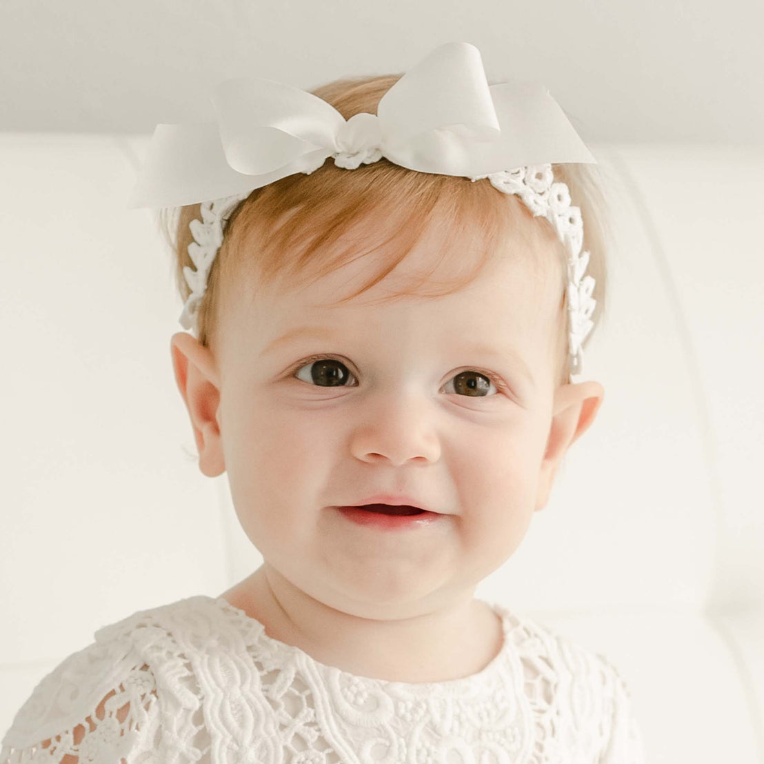 Baby girl with red hair smiling and wearing the Adeline lace headband made of cotton and silk ribbon.
