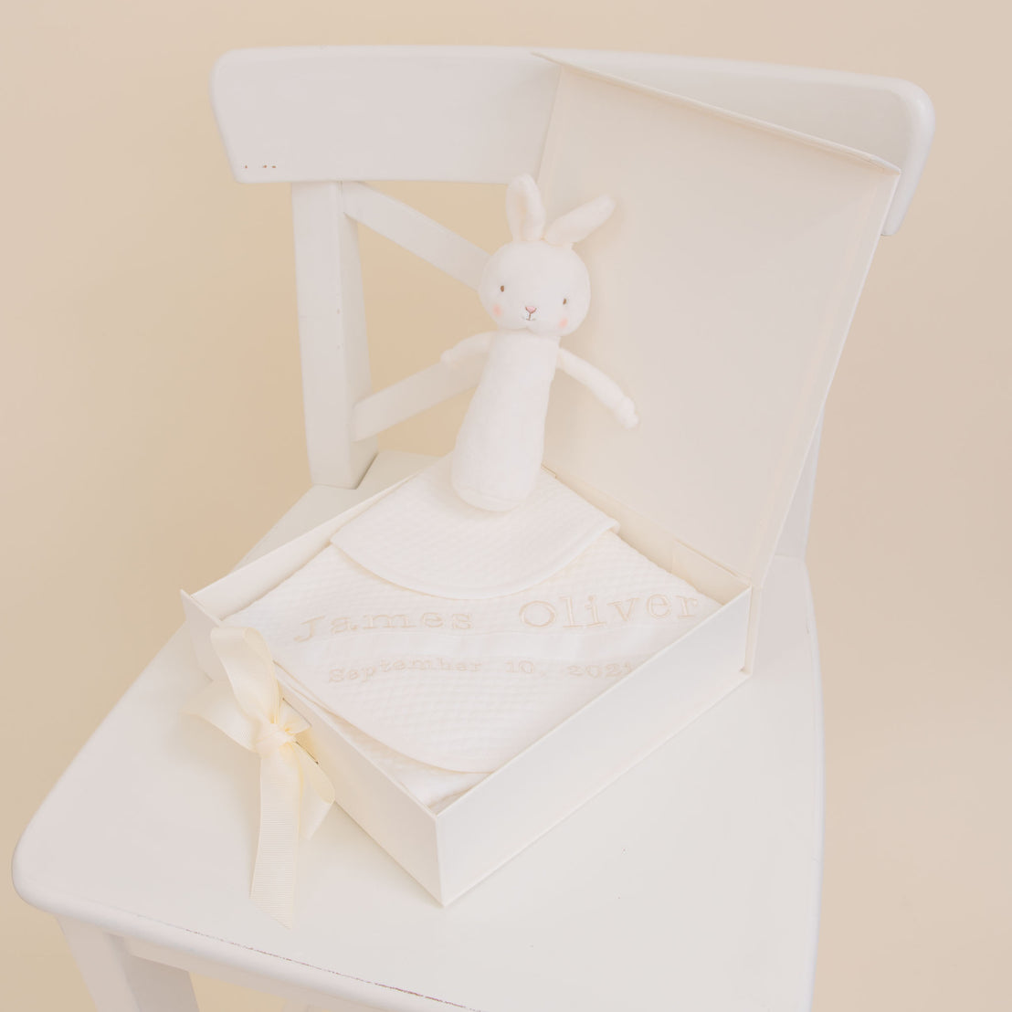 A white Baby Beau & Belle keepsake box on a chair, personalized with "James Oliver" and a date, containing an album and a stuffed rabbit. The box, an embroidered Baby Beau & Belle keepsake, is adorned with a ribbon.
