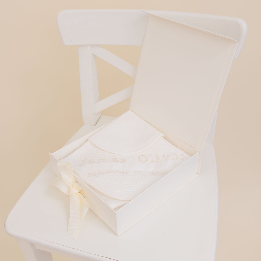 An elegant white wedding album with embroidered text "james oliver, september 10, 2023" on the cover, tied with a satin ribbon, placed on a white wooden chair against a Baby Beau & Belle Baby Boy Personalized Gift Set.