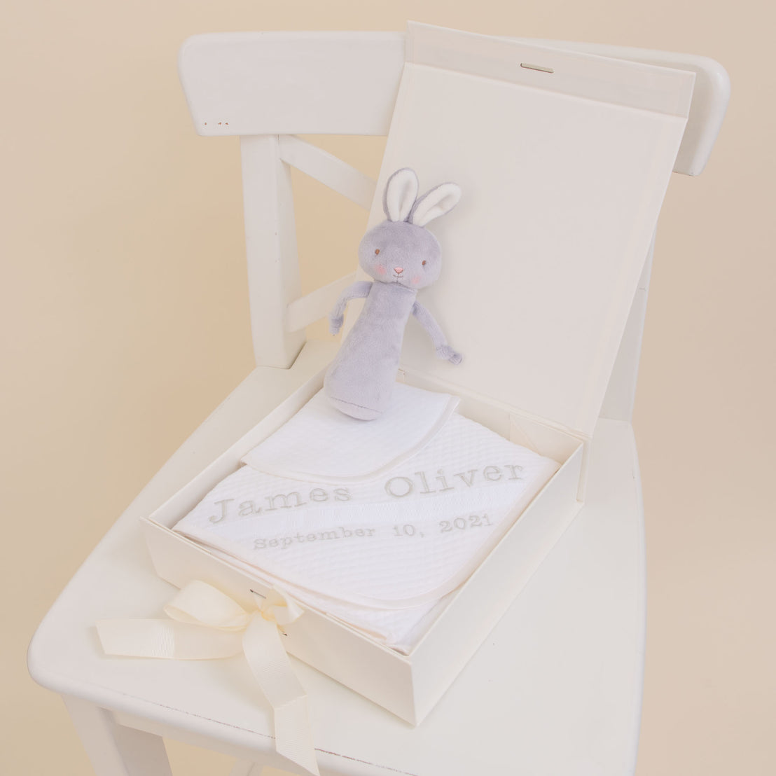 A plush bunny sits on a white chair next to an open Baby Boy Personalized Gift Set with a baby's name and date, "james oliver september 10, 2021," lined with cream by Baby Beau & Belle.