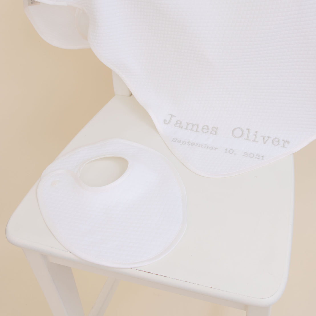 A Baby Beau & Belle quilted cotton blanket and cloth with the embroidered name "james oliver" and date "september 10, 2021", placed on a white wooden chair against a beige background