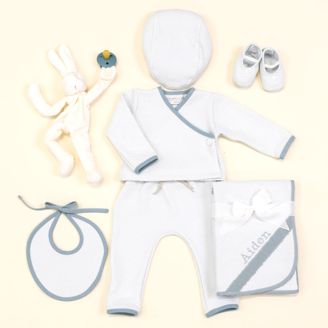 A newborn baby starter set laid out on a light background, including a cuddly bunny plush, blue and white cap, booties, bib, and the Aiden Quilted Wrap Top and Pants.