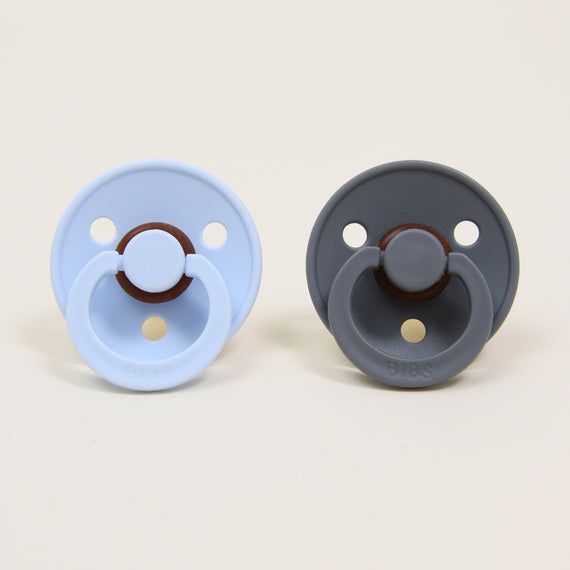 Two Bibs pacifiers on a plain background; one is in "baby blue" and the other is in "smoke", both featuring a round handle and designed as an upscale baby gift.