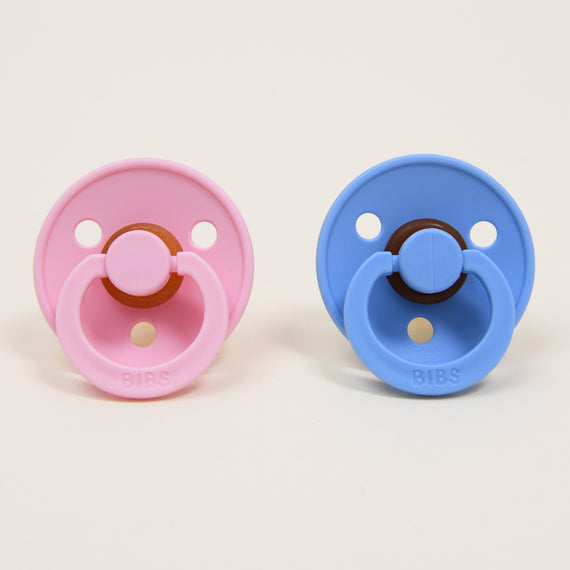 Two "Bibs" Pacifiers, one in "baby pink" and the other in "sky blue placed side by side against a beige background, each featuring a natural round button-style handle, perfect as an upscale baby gift.