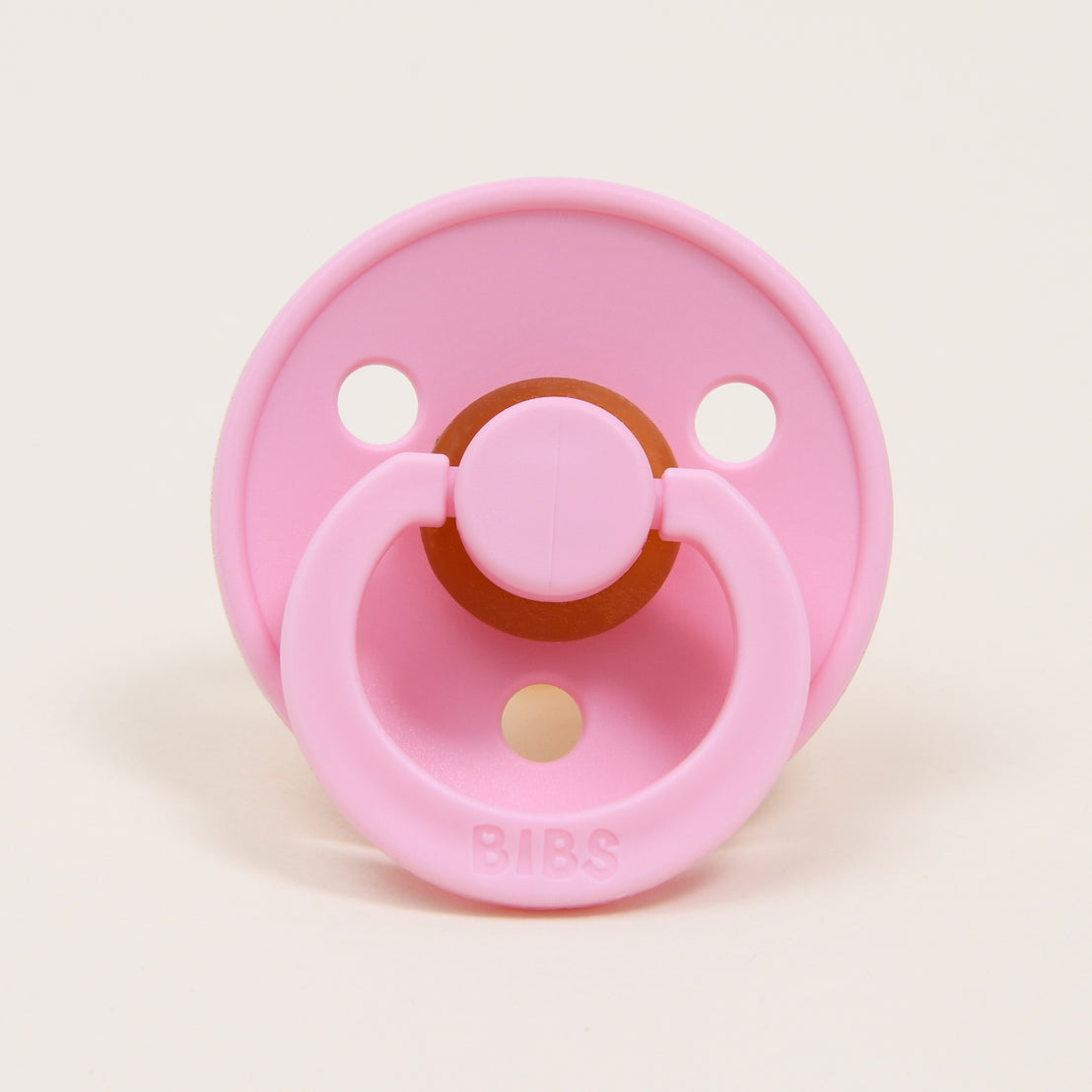 A Bibs Pacifier in Baby Pink, designed and manufactured in Denmark, with a circular rubber nipple and a round ventilation shield, labeled "bibs," on a light beige background.