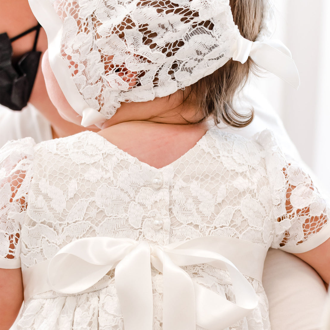 Close-up of a baby wearing the Rose Christening Gown & Bonnet, viewed from behind at a baptism. The baby is held by an adult.