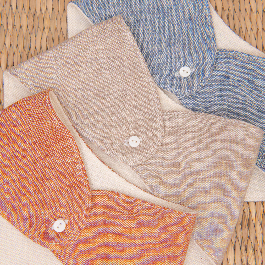 Flat lay photo of the back of all three colors of the Silas Bandana Bib. The bibs are made from linen in three different colors: indigo, sand, and clay. The back features the button closure