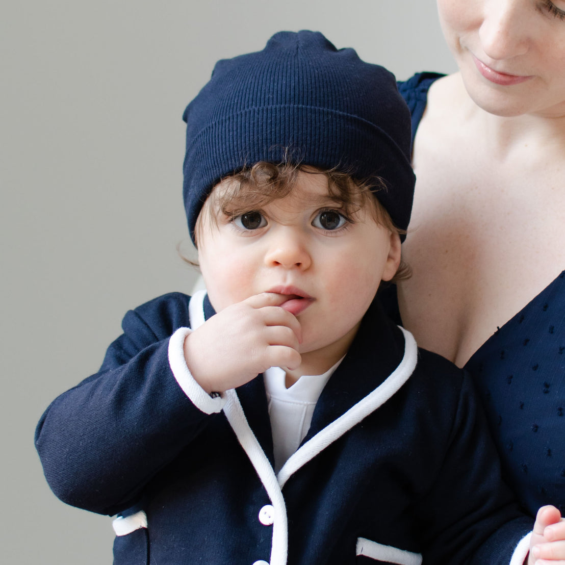 Wearing a Natural Ivory Ribbed Pima Beanie and a matching outfit with white trim, a toddler with curly hair stands next to someone in a navy top. The child gazes curiously at the camera, touching their mouth with one finger.