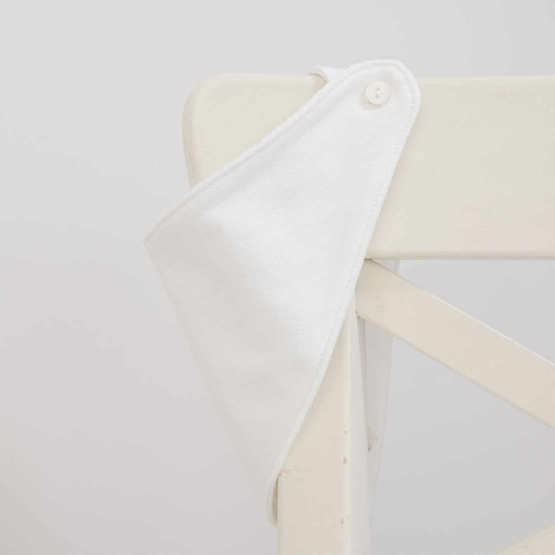 Flat lay photo of the Elliott Bib, made from white French Terry cotton, on a white chair showing the button closure.