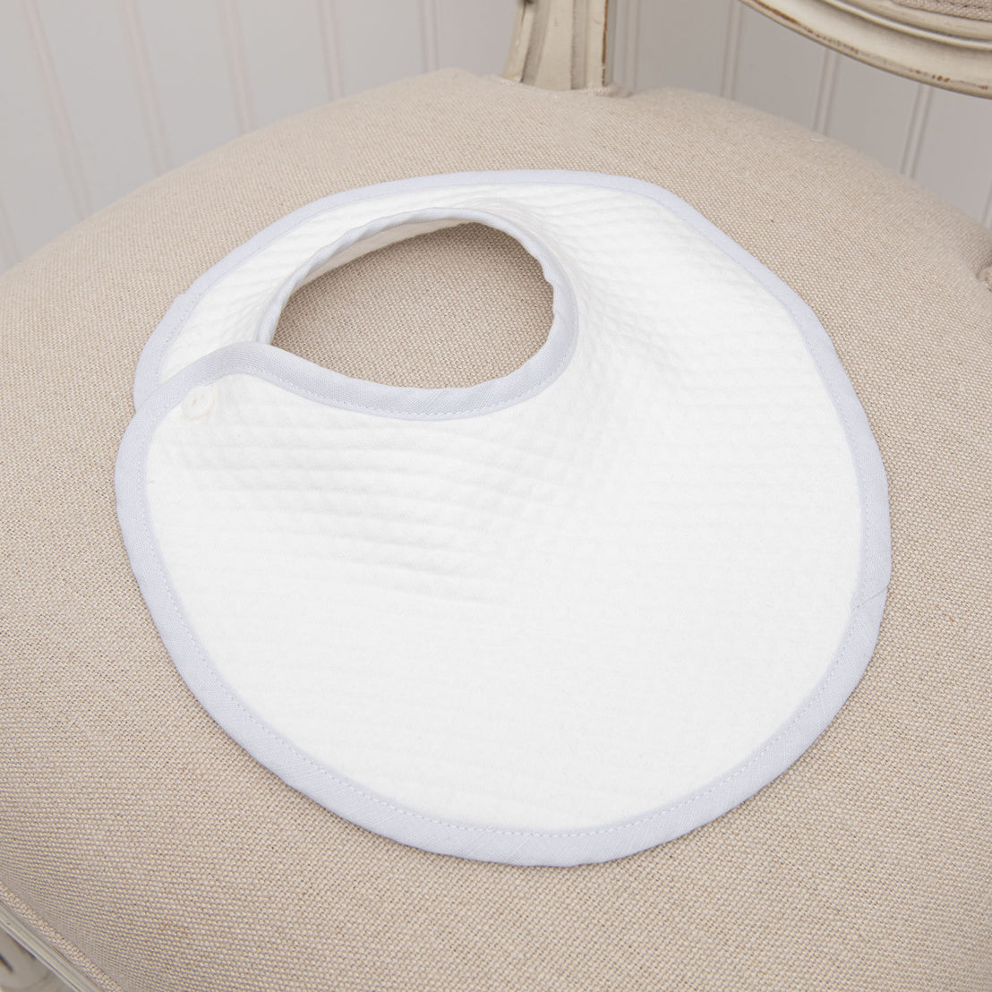 Flat of the Harrison Bib made with a very soft textured white cotton and features blue linen binding around the perimeter