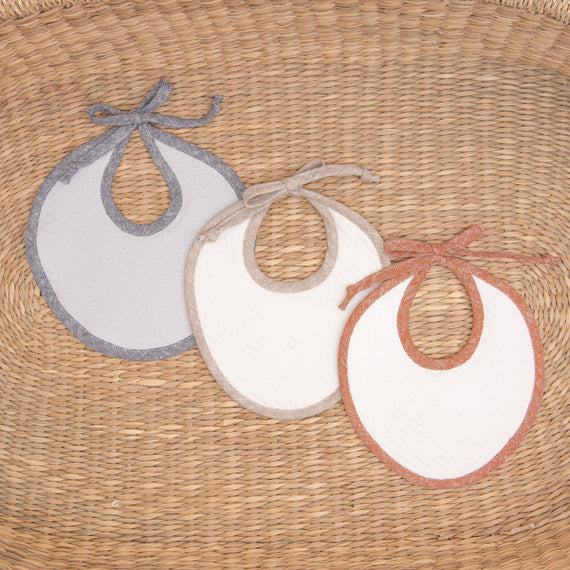 Flat lay photo of all three Silas Linen Trim Bibs, including the colors blue, tan, and red. The bibs are made from textured cotton in light ivory or grey