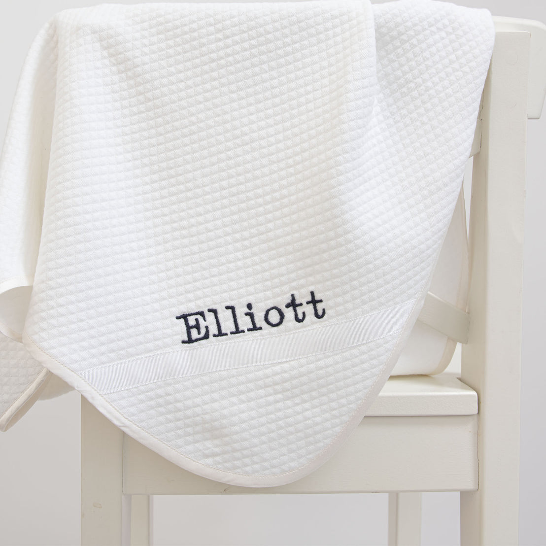 Flat lay photo of the Elliott Personalized Blanket, a soft white 100% quilted cotton, one corner is detailed with ribbon and the name "Elliott" embroidered in black..