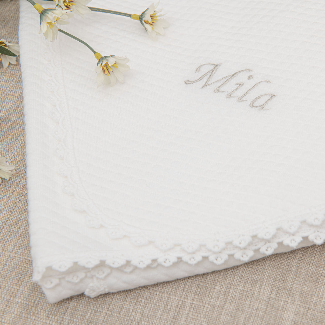 Closeup of a folded Mila Personalized Blanket with the name "Mila" embroidered in silver thread and finished with a floral edge lace on the sides. delicate floral accents, detailed lace trim. Small white flowers lie on the surface of the top left corner.