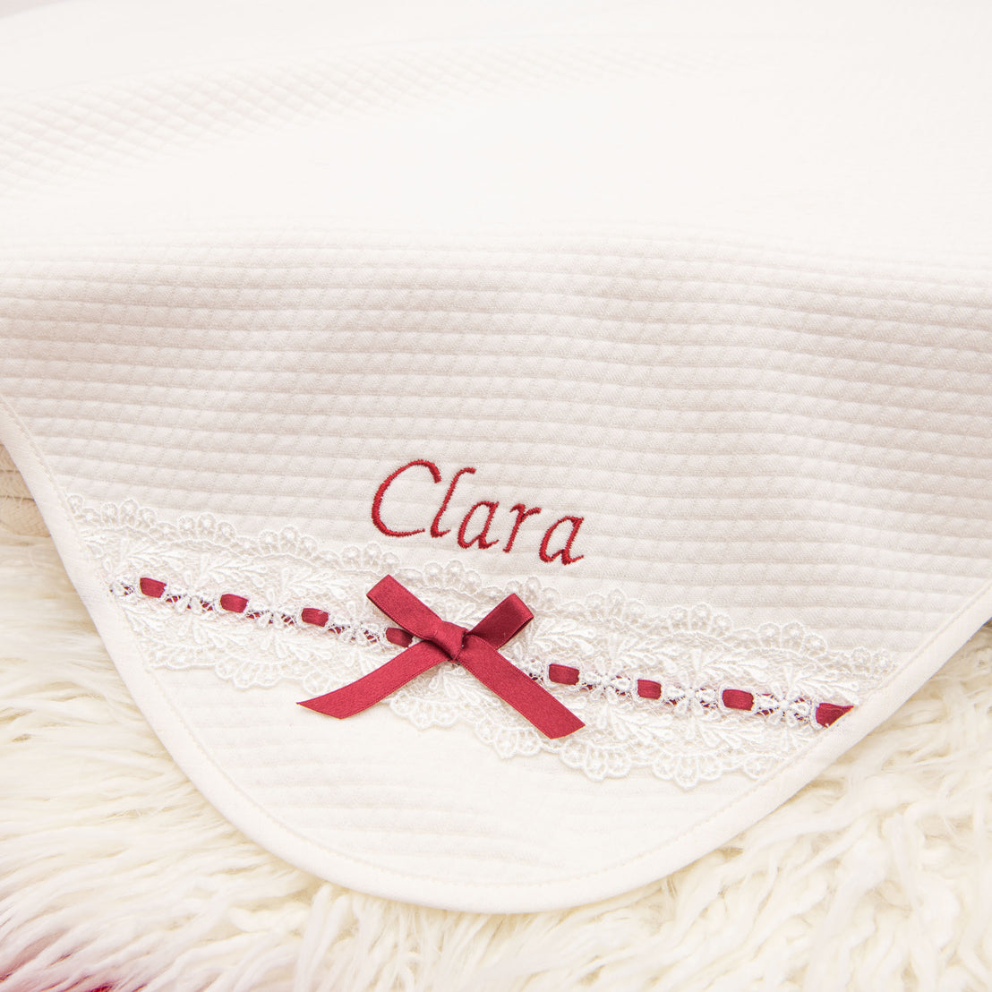 Introducing the Clara Gift Set - Save 10%: This luxurious, handmade-in-the-USA blanket features "Clara" embroidered in red on a soft white fabric. The blanket is enhanced with a beautiful lace trim adorned with a red ribbon and bow, all elegantly draped over plush white fur for an incredibly cozy and delicate look.
