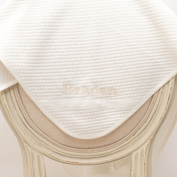Flat lay photo of the Braden Personalized Blanket made from ivory textured cotton with an ivory grosgrain ribbon woven on the corner. The name "Braden" is embroidered on the corner of the blanket.