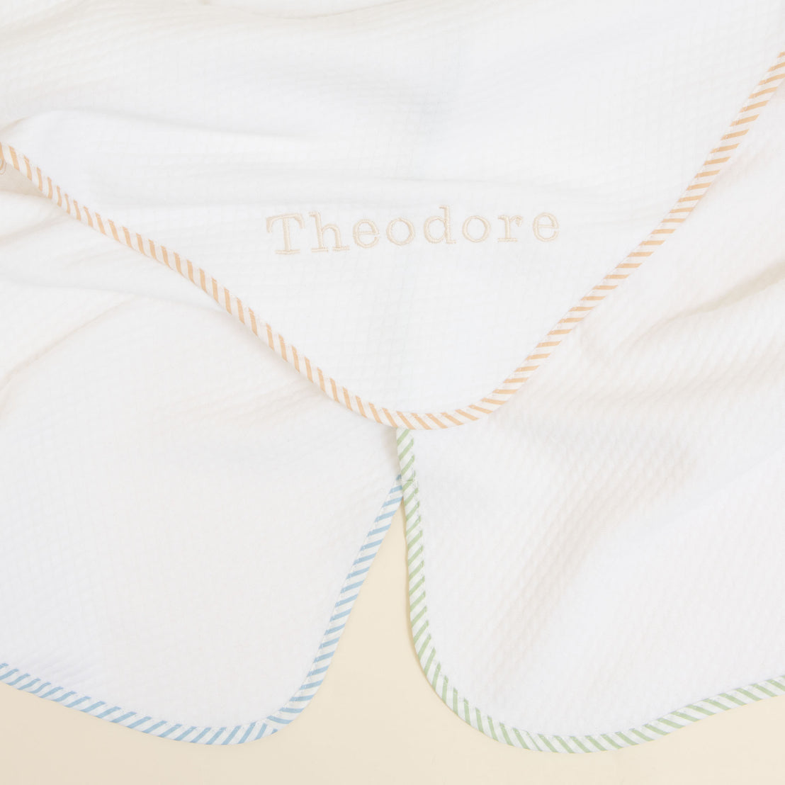 The First Easter Personalized Blanket is crafted from soft, white fabric and showcases custom embroidery of the name "Theodore" in the center. This charming gift features pastel-colored diagonal striped borders in blue, green, and orange hues, with a slightly crumpled texture adding to its appeal.