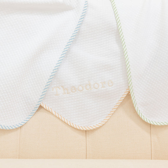 Detail of the corner of three Theodore Personalized Blankets in colors blue, green, and tan/white. Made from 100% textured cotton and featuring a ribbon corner and optional name embroidery. 