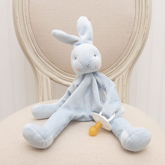 A Blue Silly Bunny Buddy | Pacifier Holder, an upscale baby gift, with a natural white nose sits on a cream upholstered chair. The bunny has long floppy ears and a serene expression.