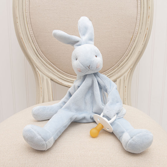 A Blue Silly Bunny Buddy | Pacifier Holder, an upscale baby gift, with a natural white nose sits on a cream upholstered chair. The bunny has long floppy ears and a serene expression.