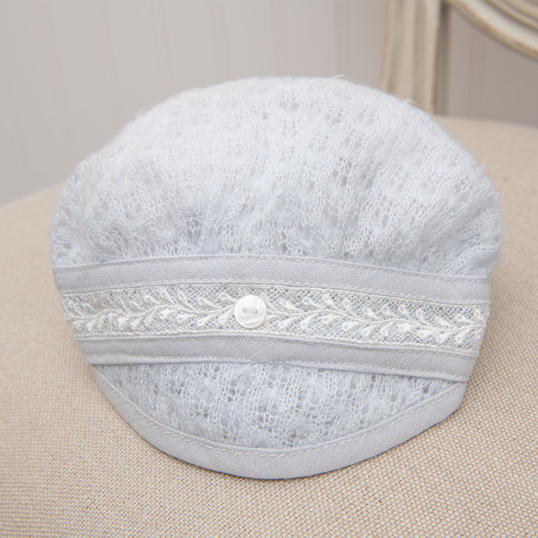 Harrison Newborn Knit Hat displayed on a neutral-toned cushion, showcasing its soft, textured knit in blue with lace trim and a button accent.