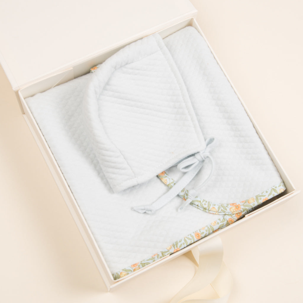 A Baby Beau & Belle gift box featuring a magnetic closure, containing a white quilted hooded towel with a floral trim, presented on a soft beige background.