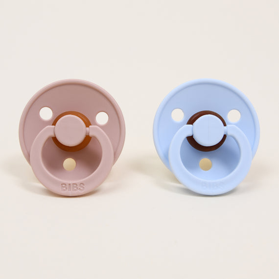 Two Bibs Pacifiers, one in blush and one in baby blue, placed side by side against a beige background, each featuring a natural round button-style handle, perfect as an upscale baby gift.