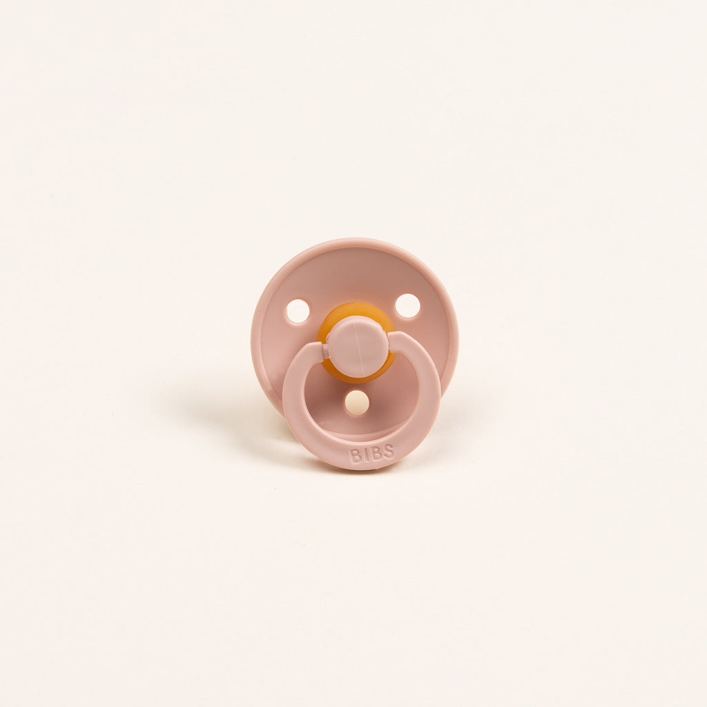 The Silly Bunny Buddy & Pacifier, in pink with a round shield and amber-colored nipple, stands upright on a white background. It features two ventilation holes and an embossed brand name on the handle. Ideal as a baby gift, this delightful accessory pairs perfectly with any pacifier holder for added convenience.