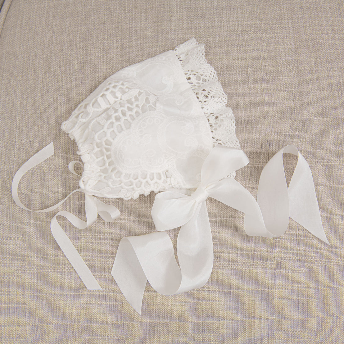 Flat lay of the Lily Bonnet on a beige fabric surface, highlighting the light ivory cotton lace details, edge lace, and ivory silk ribbons.