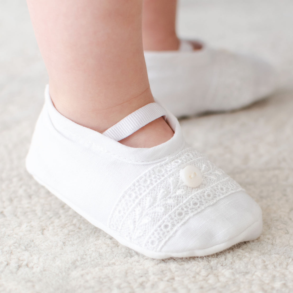 The top detail of the Oliver Booties in white worn by a baby boy. The Oliver booties are made with a linen top, soft pima cotton bottom, and features a Venice lace and button detail (with elastic strap across the foot).