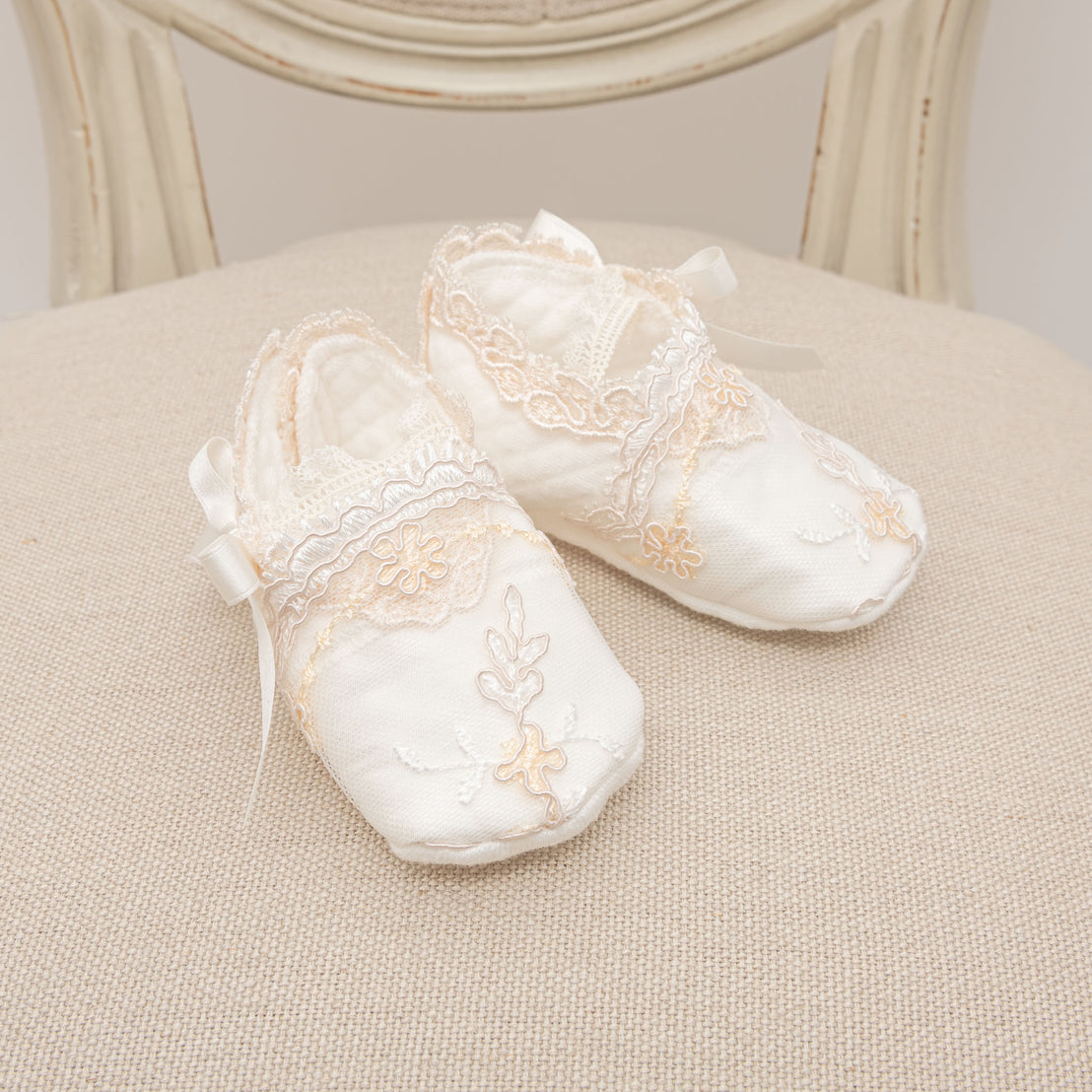 A pair of delicate, white Kristina baby shoes with floral patterns and lace details, resting on a textured beige cushion against a vintage-inspired chair background.