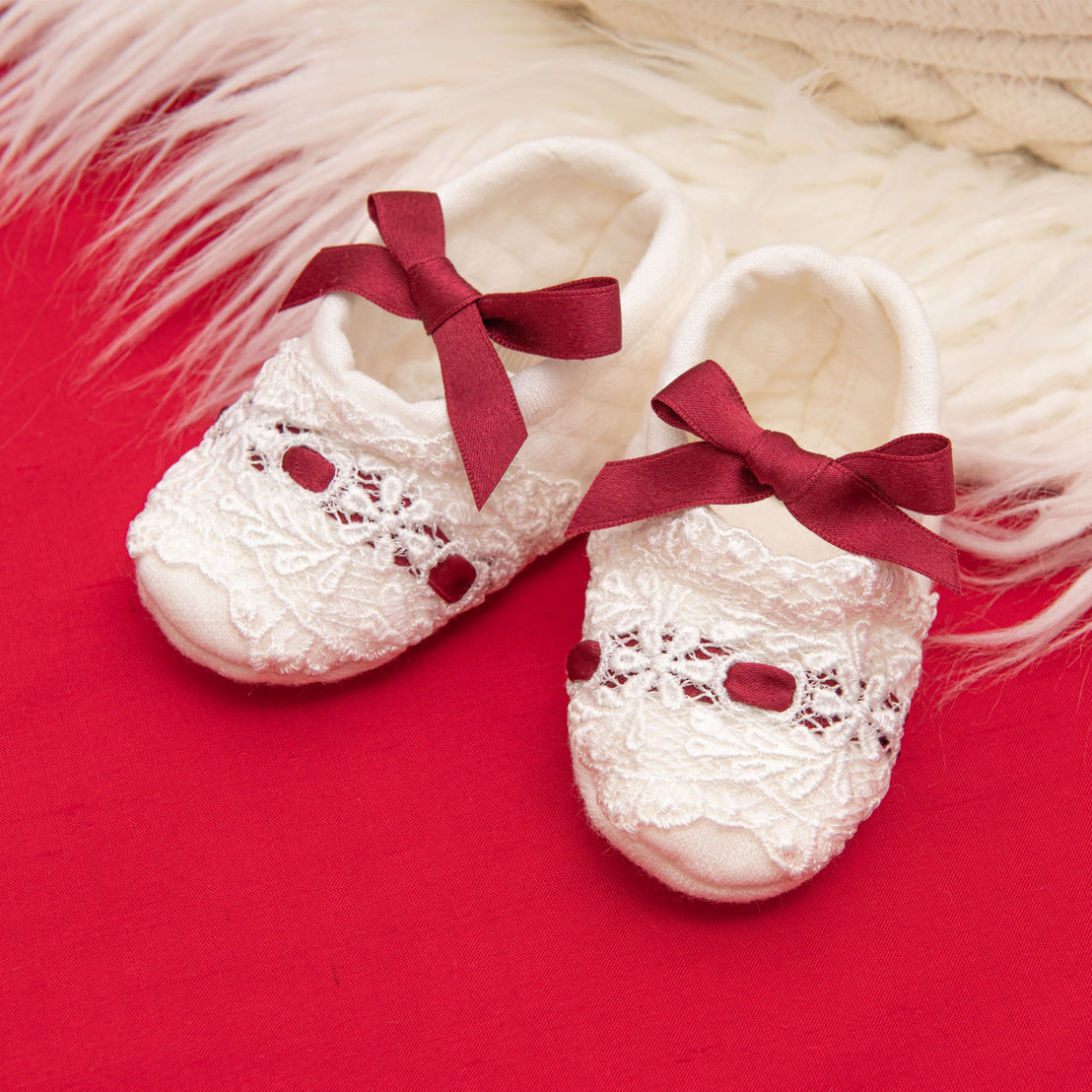 The Clara Quilted Booties, featuring lace detailing and maroon silk ribbon bows on top, rest on a plush white surface against a red fabric backdrop.