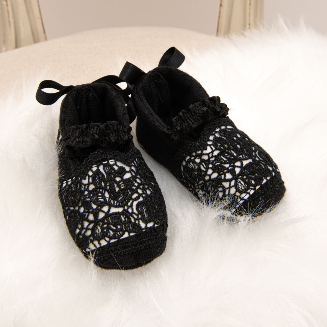A pair of June Black Lace Booties with ribbon ties, positioned on a soft white fur surface, perfect as a baby shower gift.