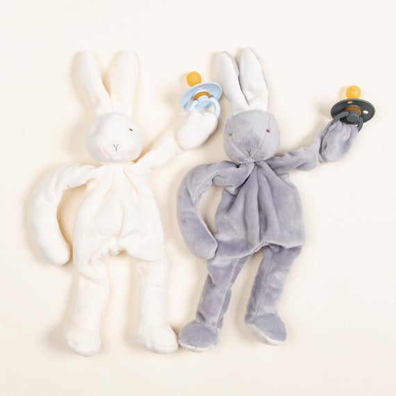 The Silly Bunny Buddy & Pacifier set includes two plush bunny toys, perfect for a baby gift, with one in cream and the other in gray. Each cuddly bunny holds a pacifier, features long ears and simple embroidered faces on their soft bodies, and they are arranged side by side on a light background.