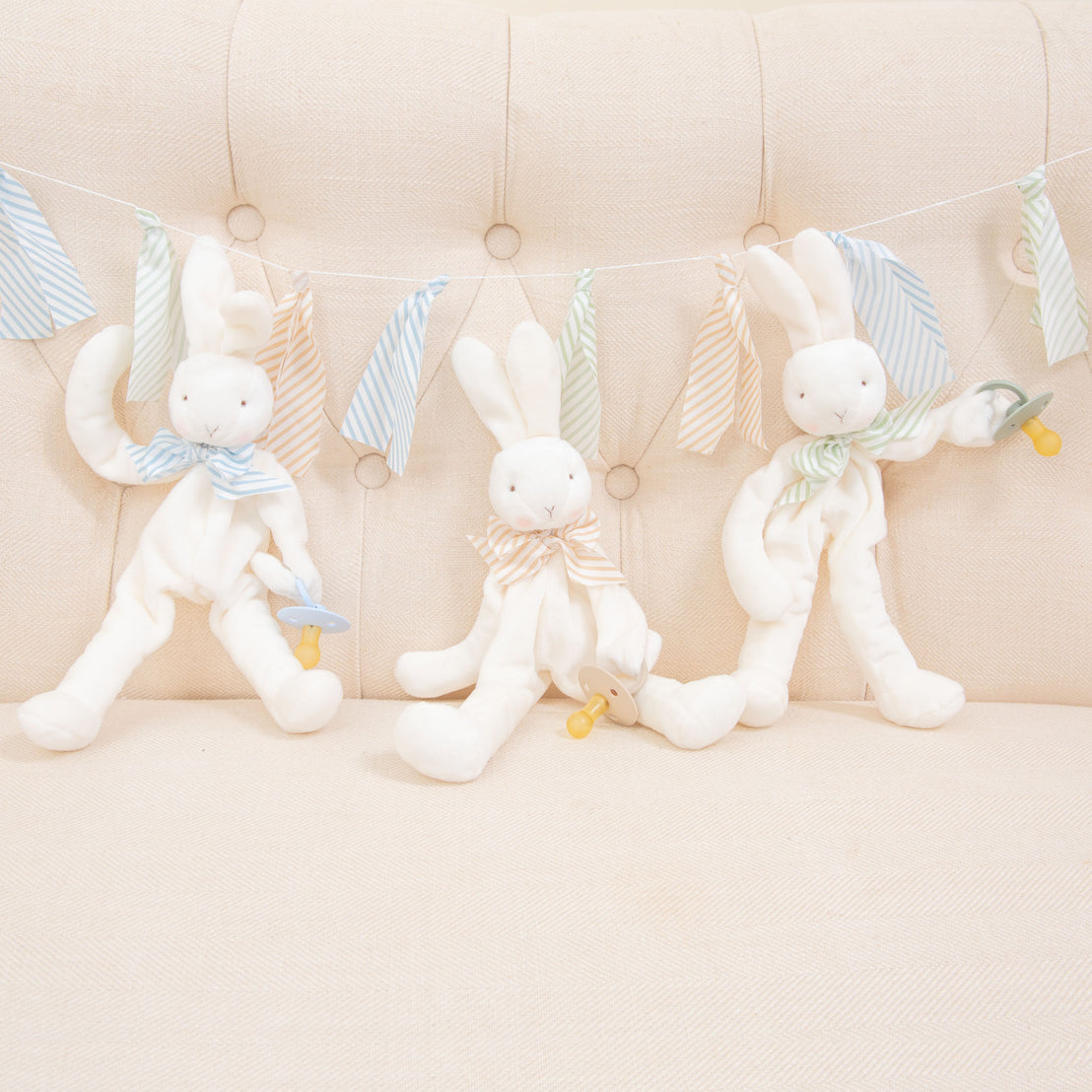 Flat lay photo of three Theodore Bunny Buddy Pacifier Holders. Stuffed animal floppy bunny made from a soft velour. Available colors include blue, green, and tan/white.