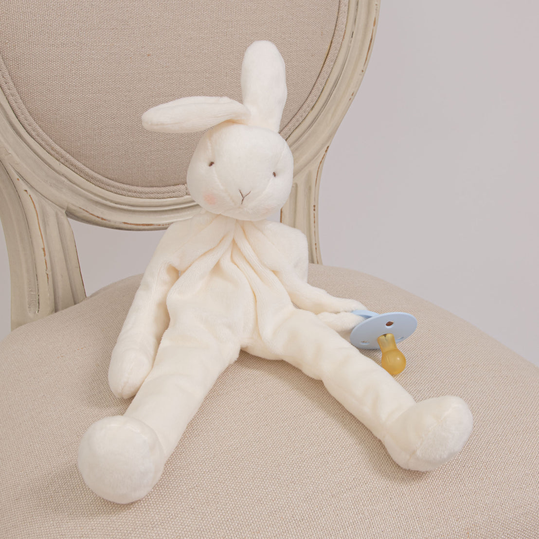 The Owen Silly Bunny Buddy and Pacifier sitting on a chair. It is a stuffed bunny holding a baby blue pacifier.