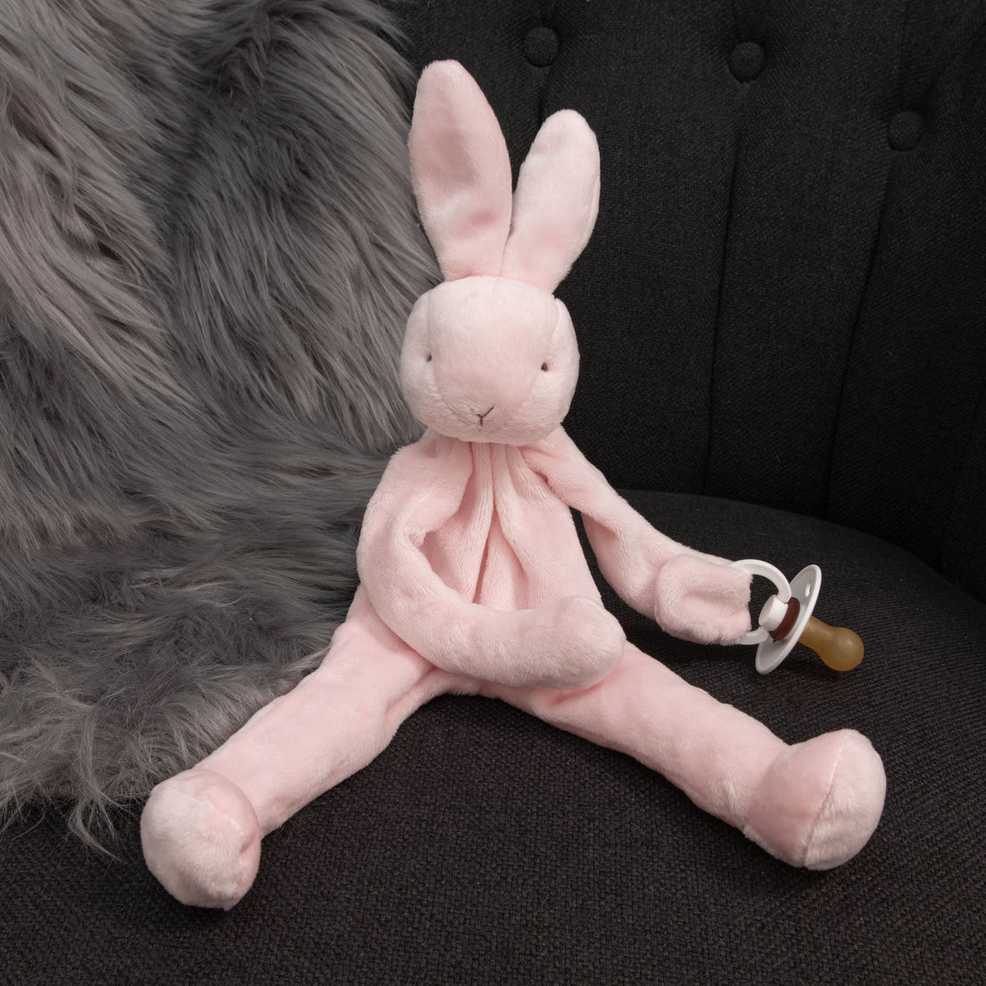 A Pink Silly Bunny Buddy, designed as a pacifier holder, rests on a dark cushioned chair. This charming baby gift gently holds a pacifier, with a fluffy gray fur blanket partially draped behind it.