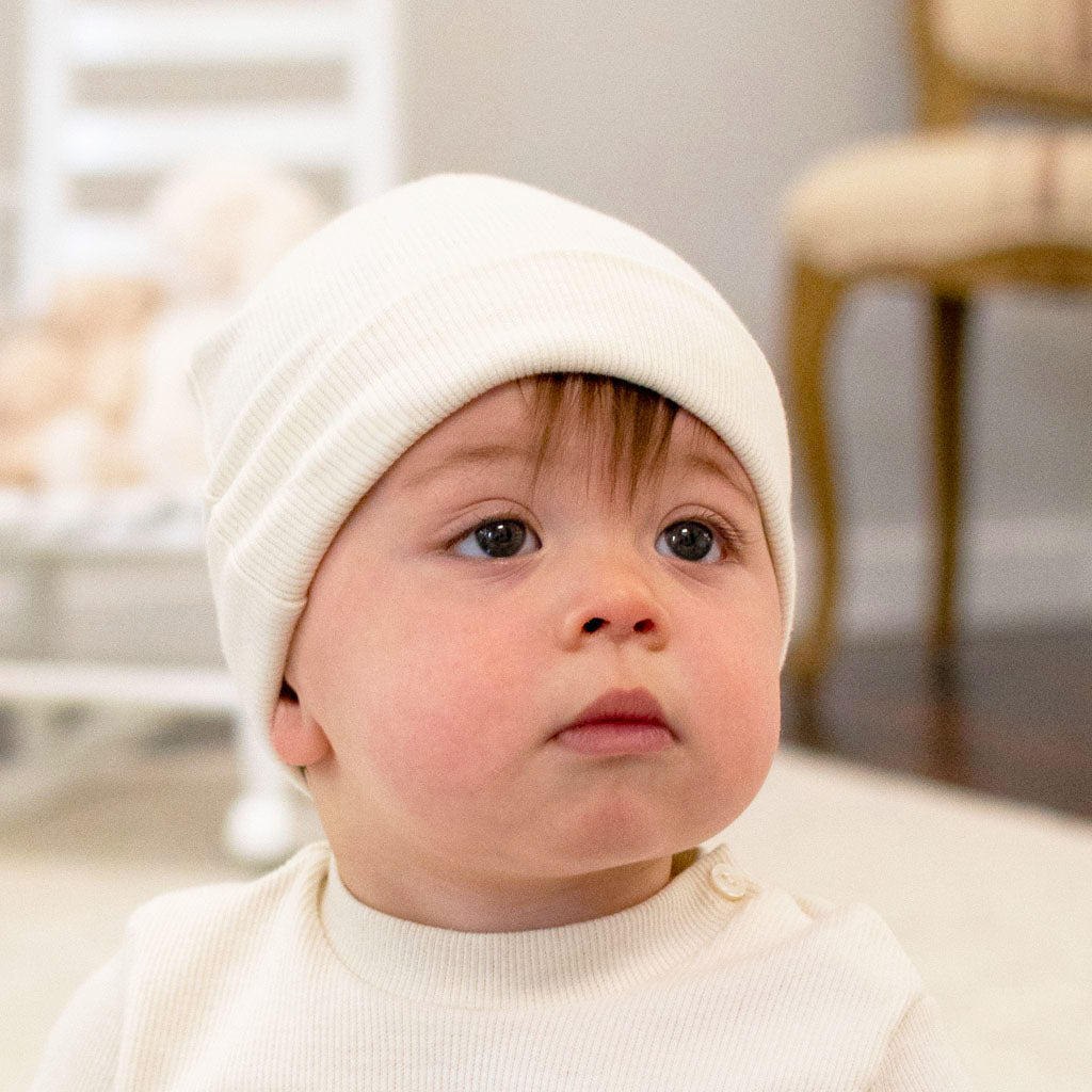 Wearing a Natural Ivory Ribbed Pima Beanie and a light-colored shirt, the baby gazes upward with a curious expression. The background showcases a blurred chair and crib, adding to the cozy indoor ambiance.