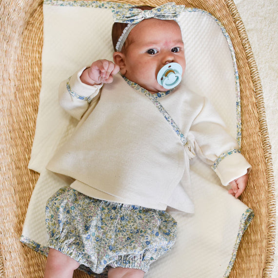 A baby with a pacifier lies in a vintage-inspired wicker basket, dressed in a beige top and Petite Fleur Bloomers, with a matching headband.