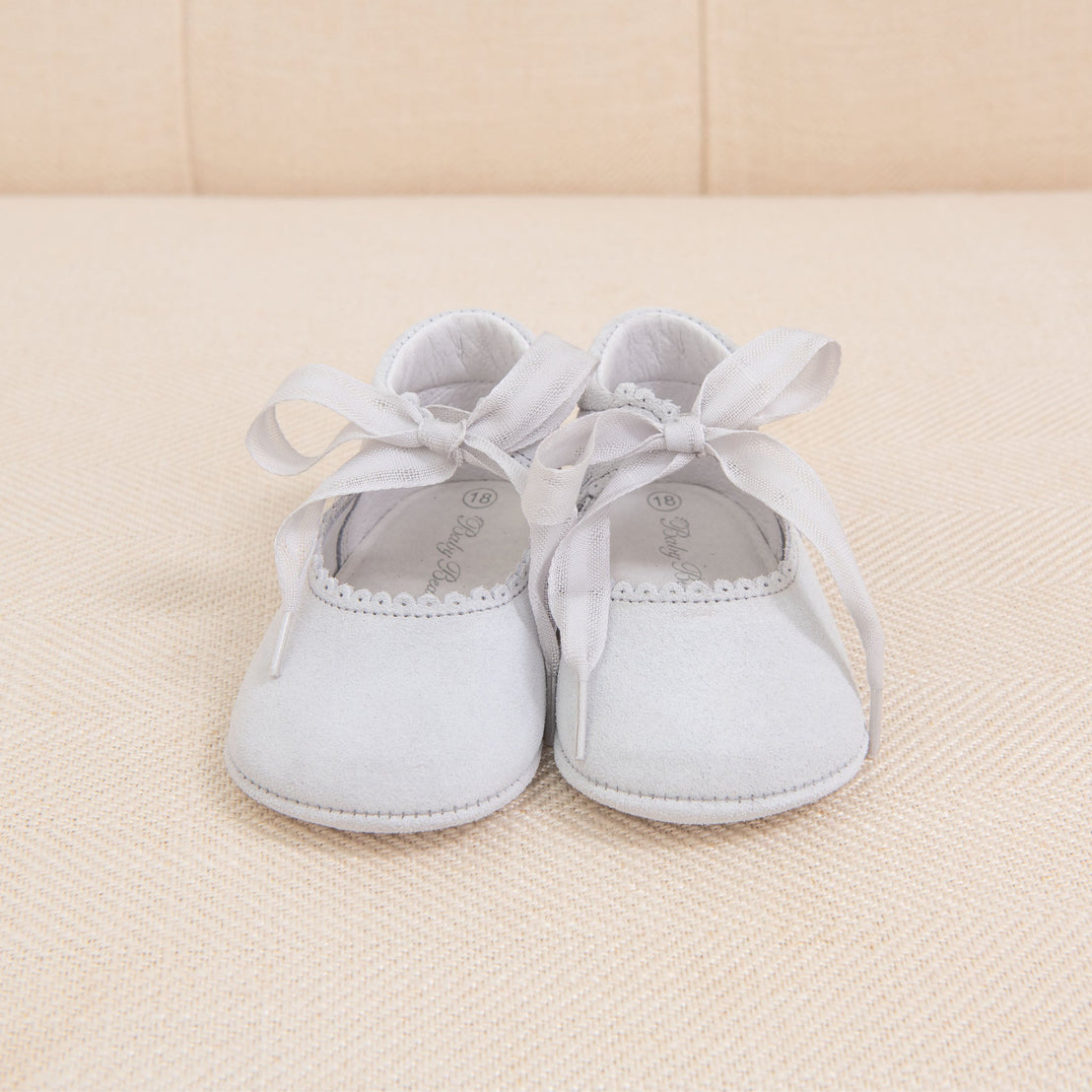 A pair of Thea Suede Tie Mary Janes in light gray with white ribbon ties, ideal for a first birthday, placed on a traditional beige fabric surface.