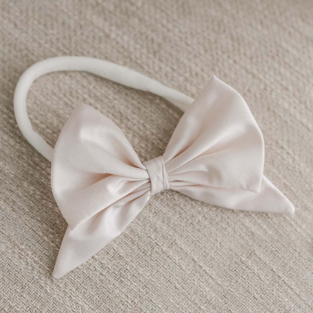 A Elizabeth Silk Bow Headband placed on a textured beige fabric, neatly tied and centered on the band, presenting an elegant and upscale appearance suitable for a baptism.