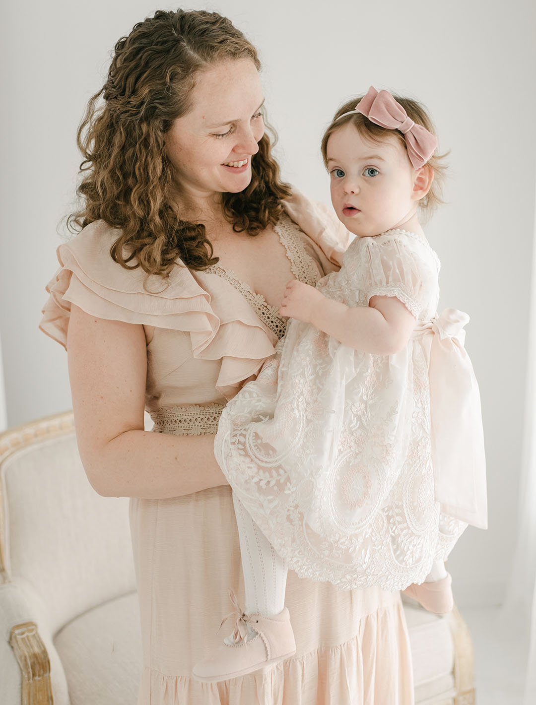 A woman with curly hair in a ruffled dress cradles a young child wearing the Elizabeth Dress, featuring embroidered lace and a delicate bow headband. They are in a softly lit room with light-colored furniture, both looking content.