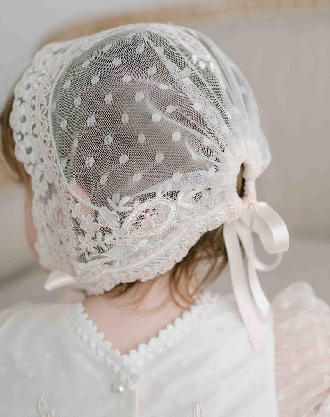Back view of a baby girl wearing the Elizabeth Lace Bonnet and matching dress. The angle showcases the pink dotted mesh embroidered lace and cinched silk ribbon ties.