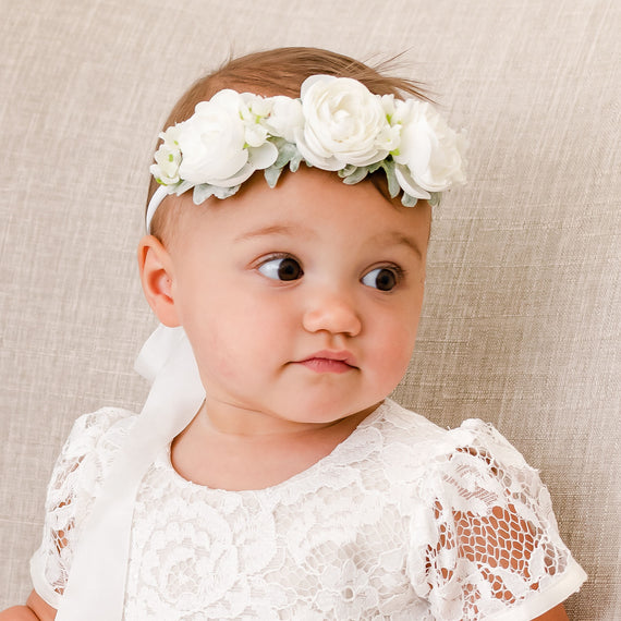 A baby dressed in a white lace dress sits on a beige couch, wearing the Rose Flower Headband made from hand-stretched nylon. The headband, adorned with ivory roses, beautifully frames her face as she gazes to the side with a curious expression.