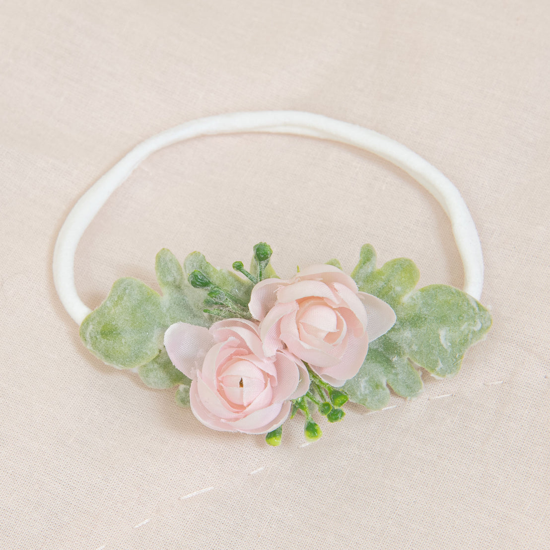 The Natalie Flower Headband showcases two pink roses and green leaves, beautifully arranged on a light beige fabric background, skillfully crafted from hand-stretched nylon.